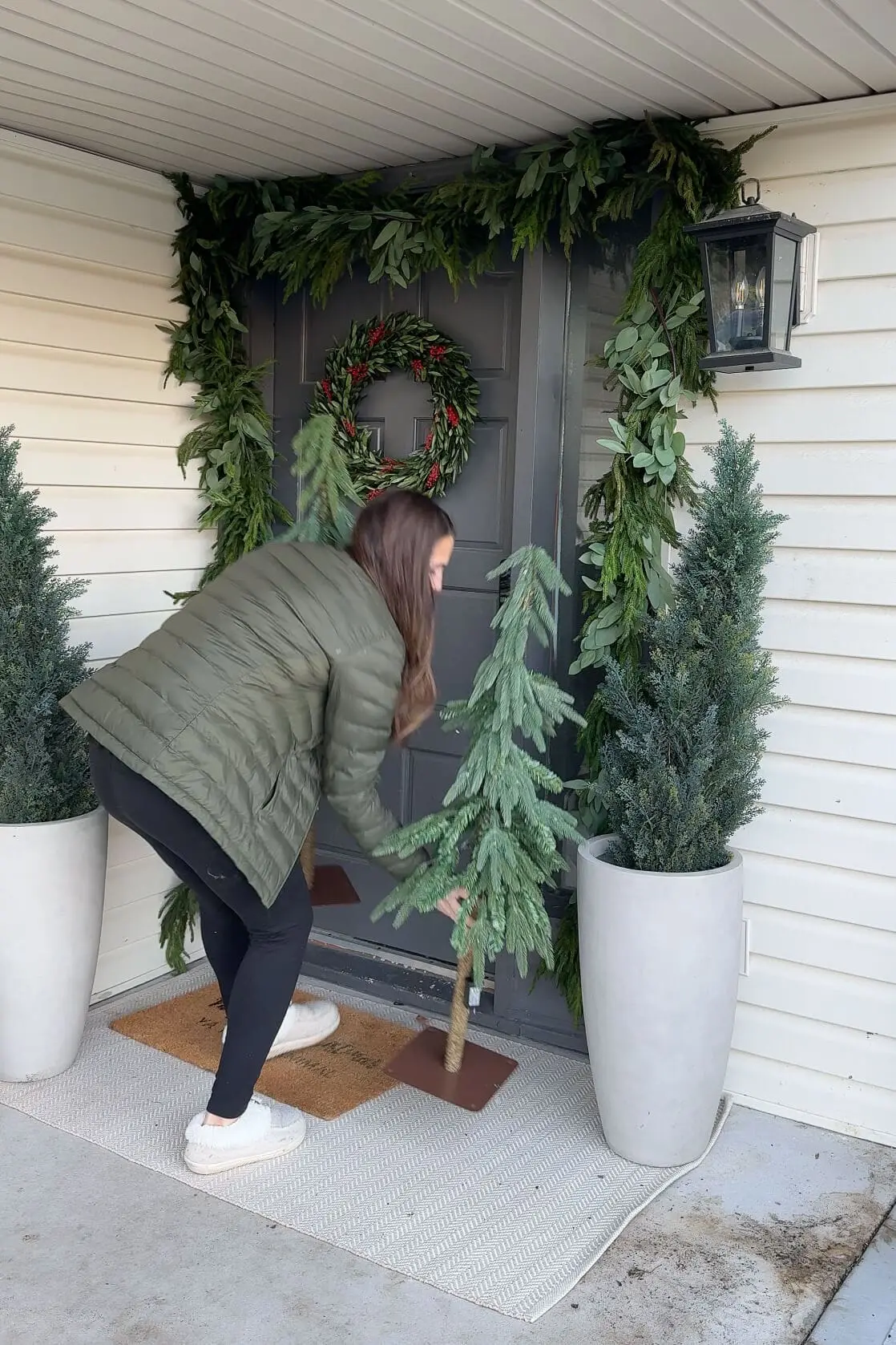 A woman setting up small artificial Christmas trees next to tall potted evergreens on a front porch, beautifully adorned with fresh greenery garland and a holiday wreath on a gray door.